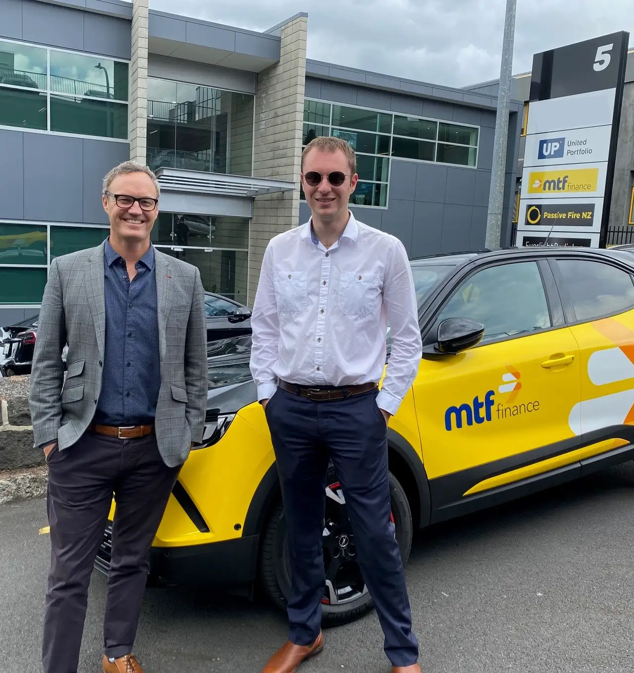 Two men standing in front of a yellow MTF Finance branded car